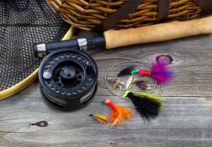 Close up top view of fishing fly reel, landing net, creel and assorted flies, with partial cork handled pole on rustic wooden boards